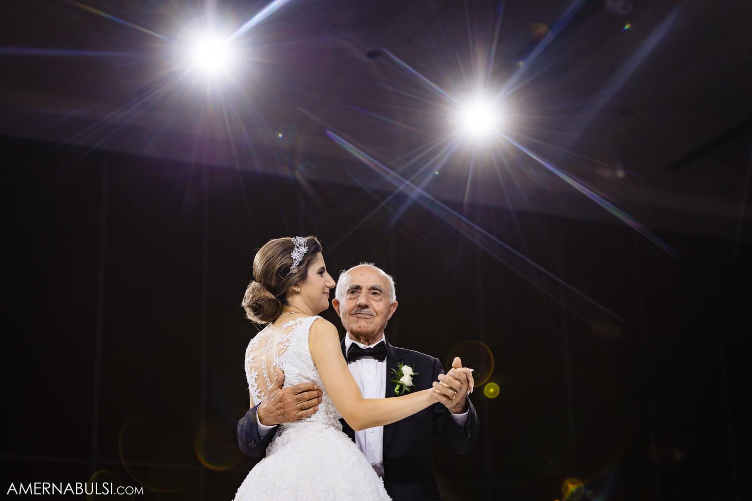 A couple shares their first dance as bright spotlights illuminate them in an elegant ballroom setting.