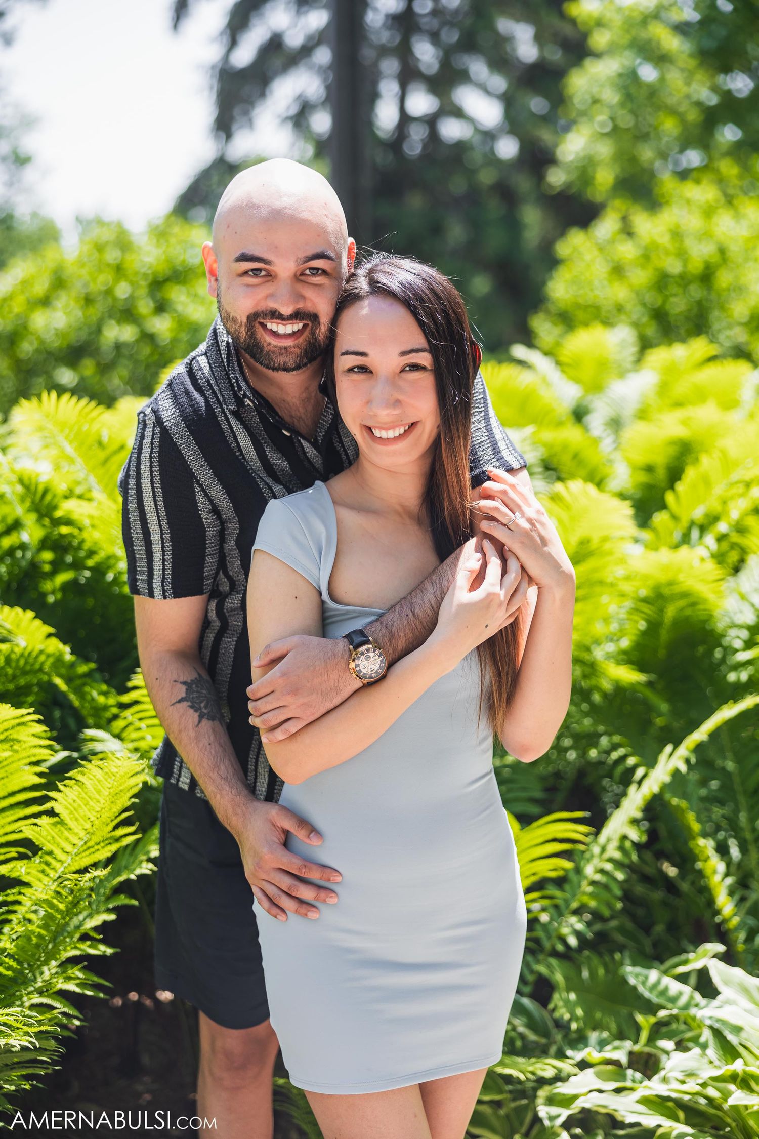 A happy couple poses together in front of green ferns on a bright summer day.