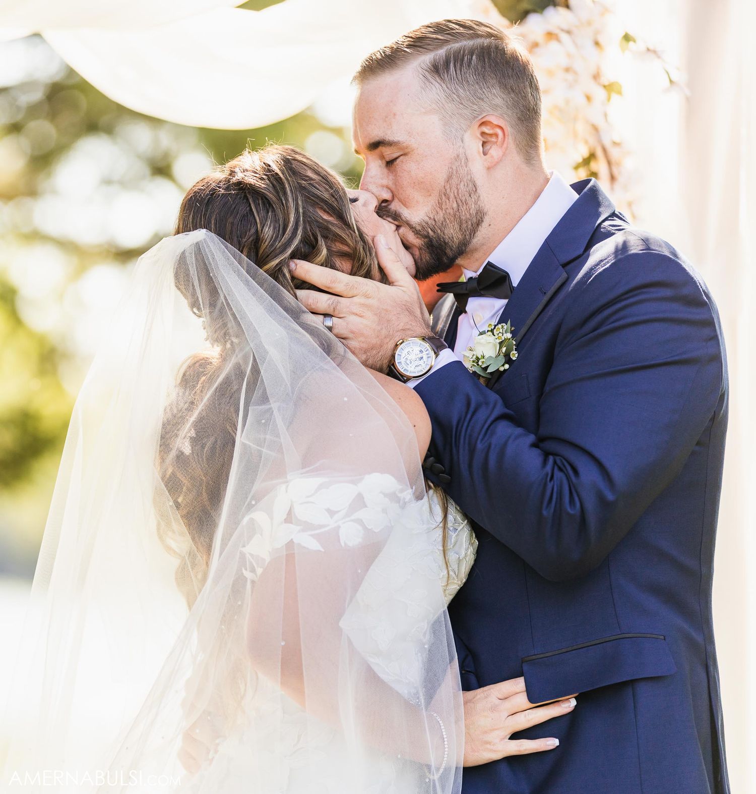 A tender kiss shared between newlyweds under flowing white veils during their outdoor ceremony.
