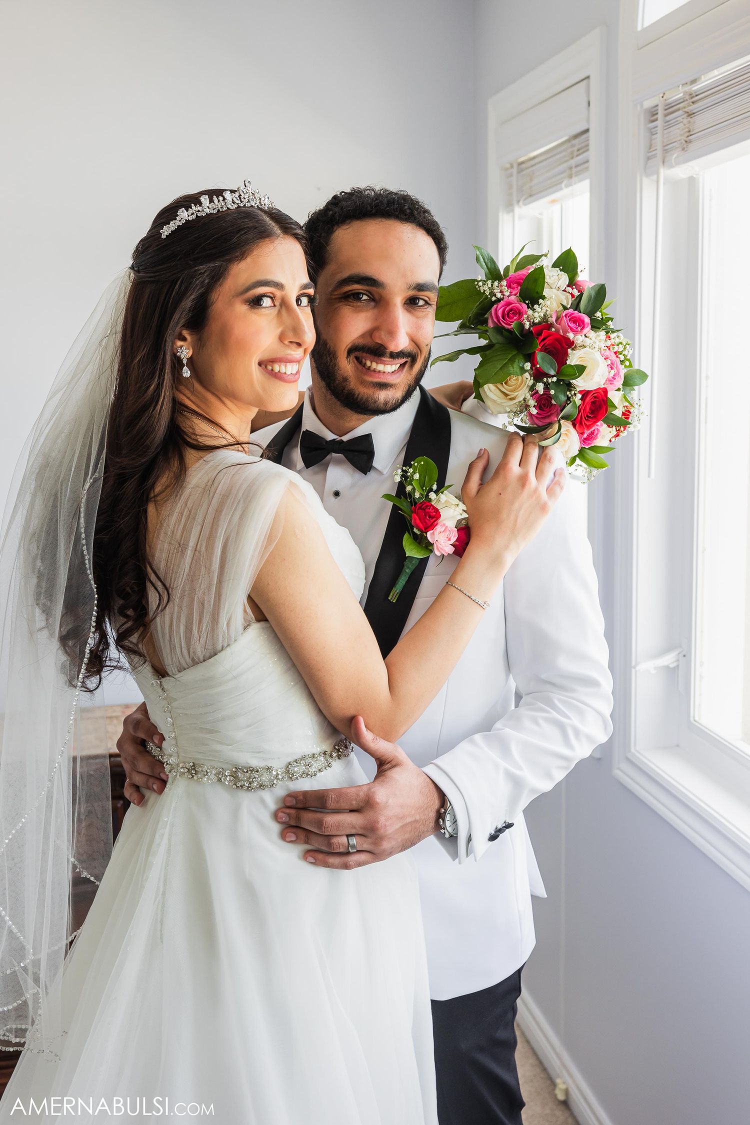 A couple poses near a bright window with a vibrant pink and red bouquet.