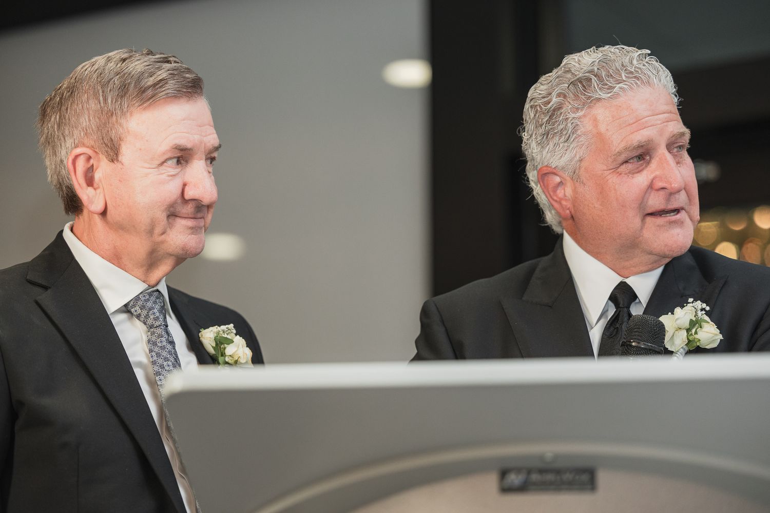 Two men in black suits with white boutonnieres smiling at a formal wedding ceremony.