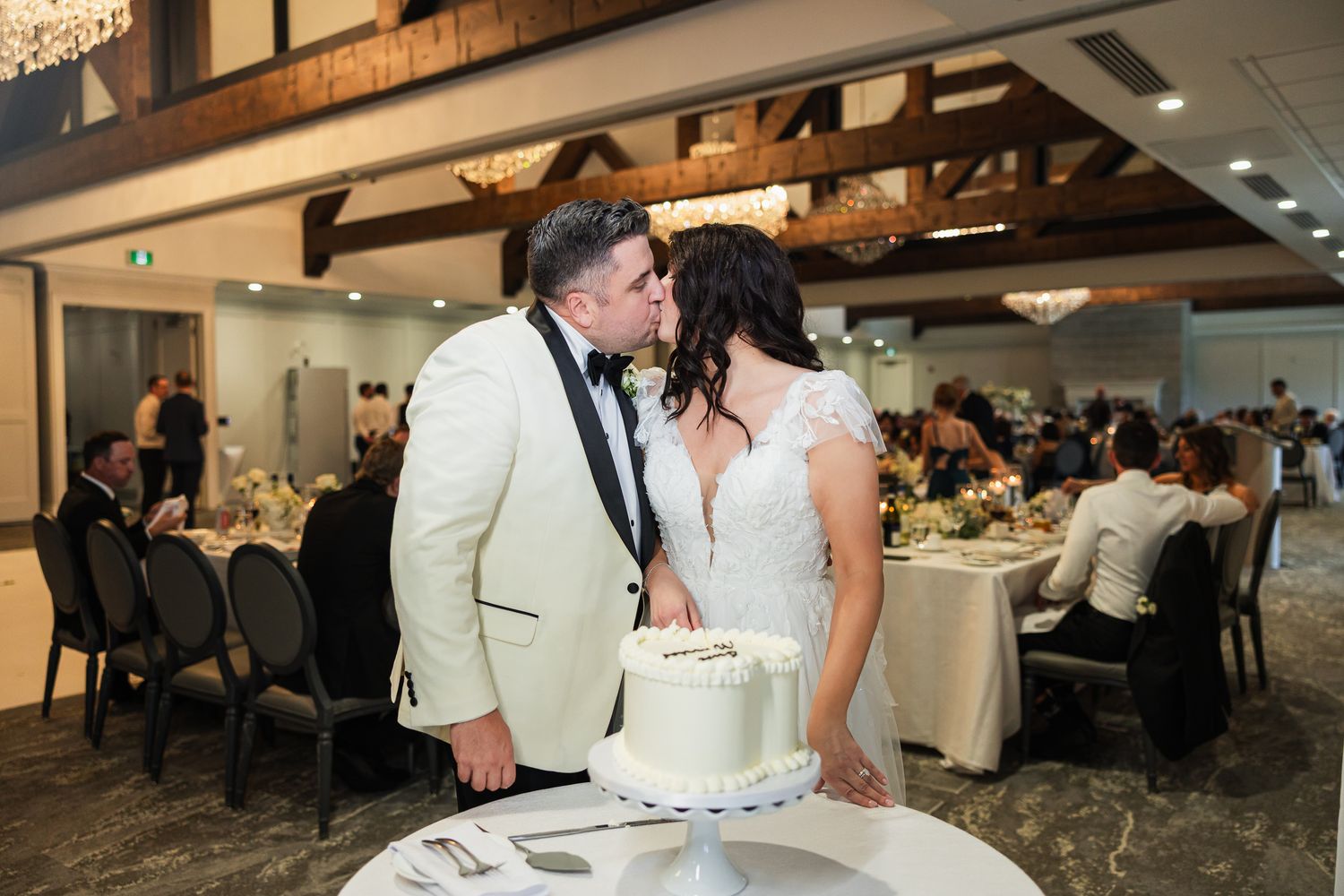 Newlyweds share a romantic kiss while cutting their white wedding cake at reception.