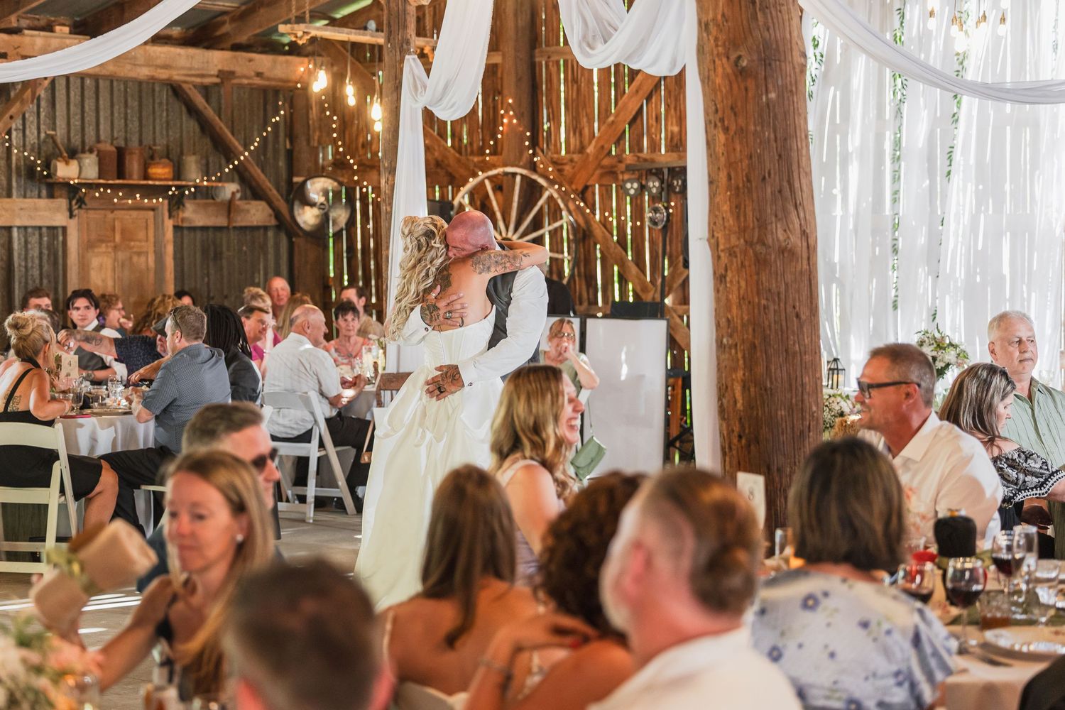 Bride and groom share first dance in a rustic barn wedding venue decorated with string lights and white draping.