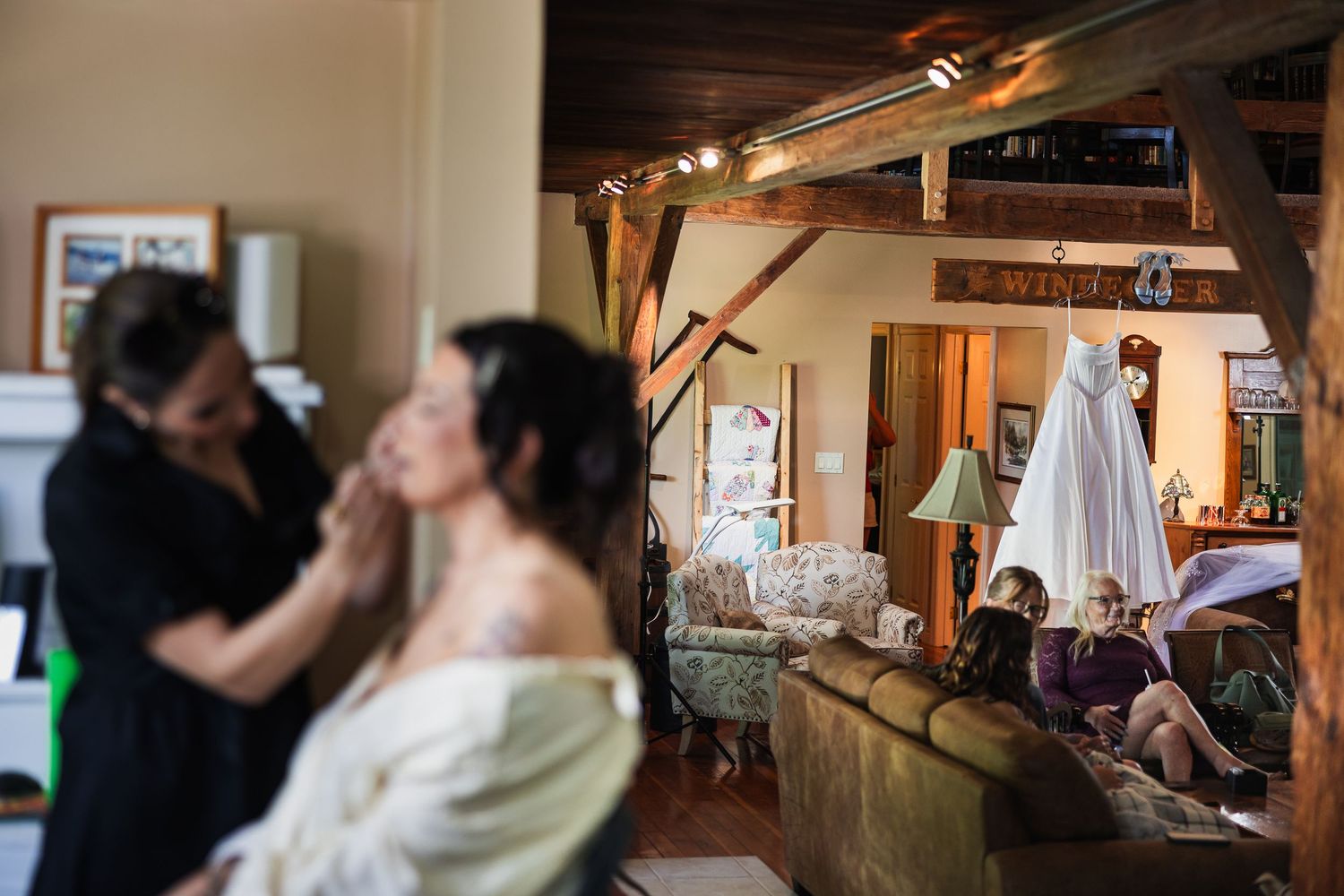 Bride getting makeup done in rustic venue with wedding dress hanging in background.