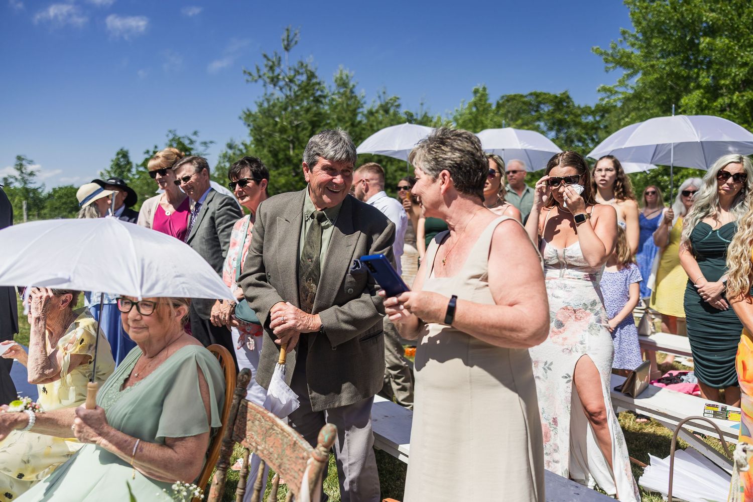 Wedding guests using white umbrellas for shade at sunny outdoor ceremony while couple exchanges rings.