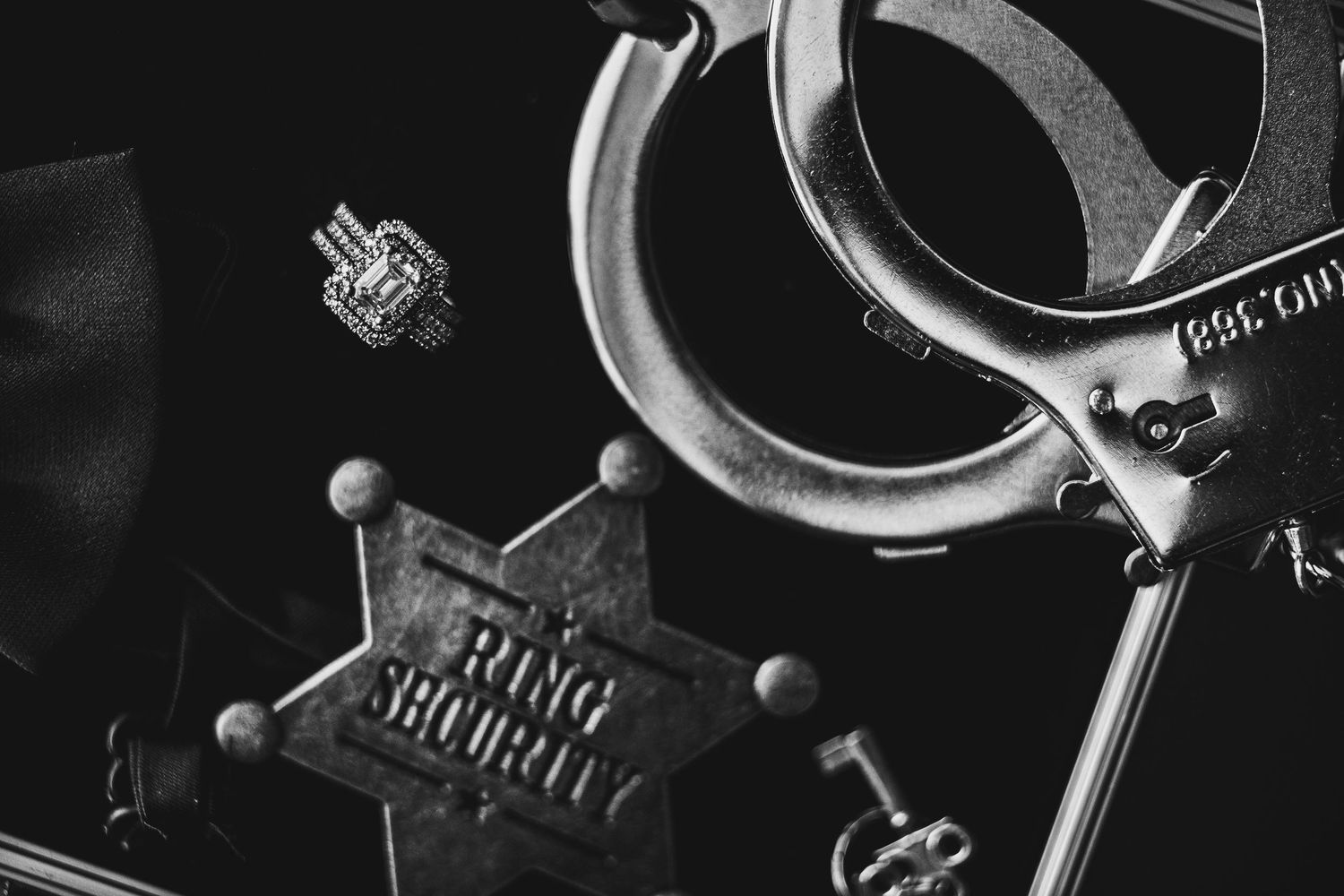 Black and white photo of ring security badge, handcuffs, and wedding ring on dark surface.