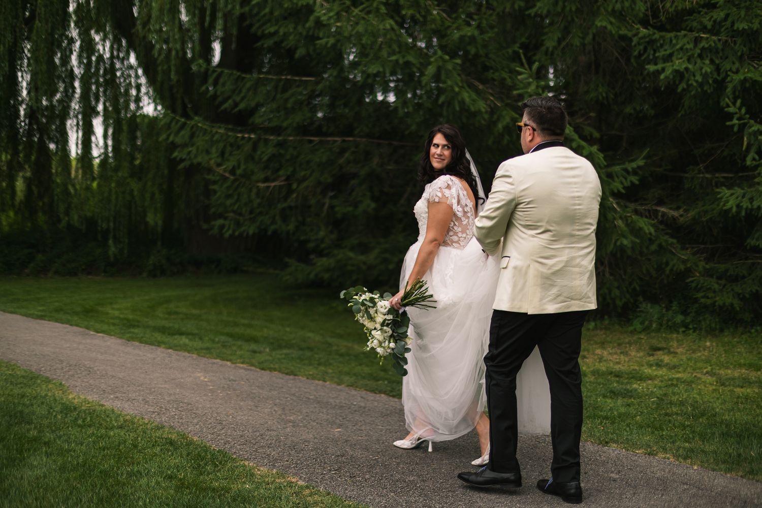 Bride holding white bouquet glances back at groom on a tree-lined garden path at their wedding.