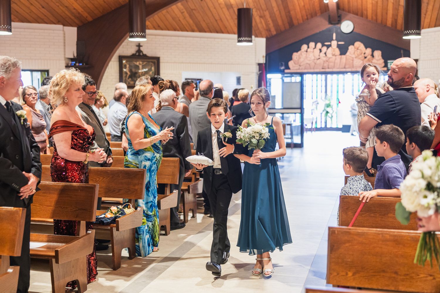 Wedding party members walk down the church aisle during a ceremony with guests seated in pews.