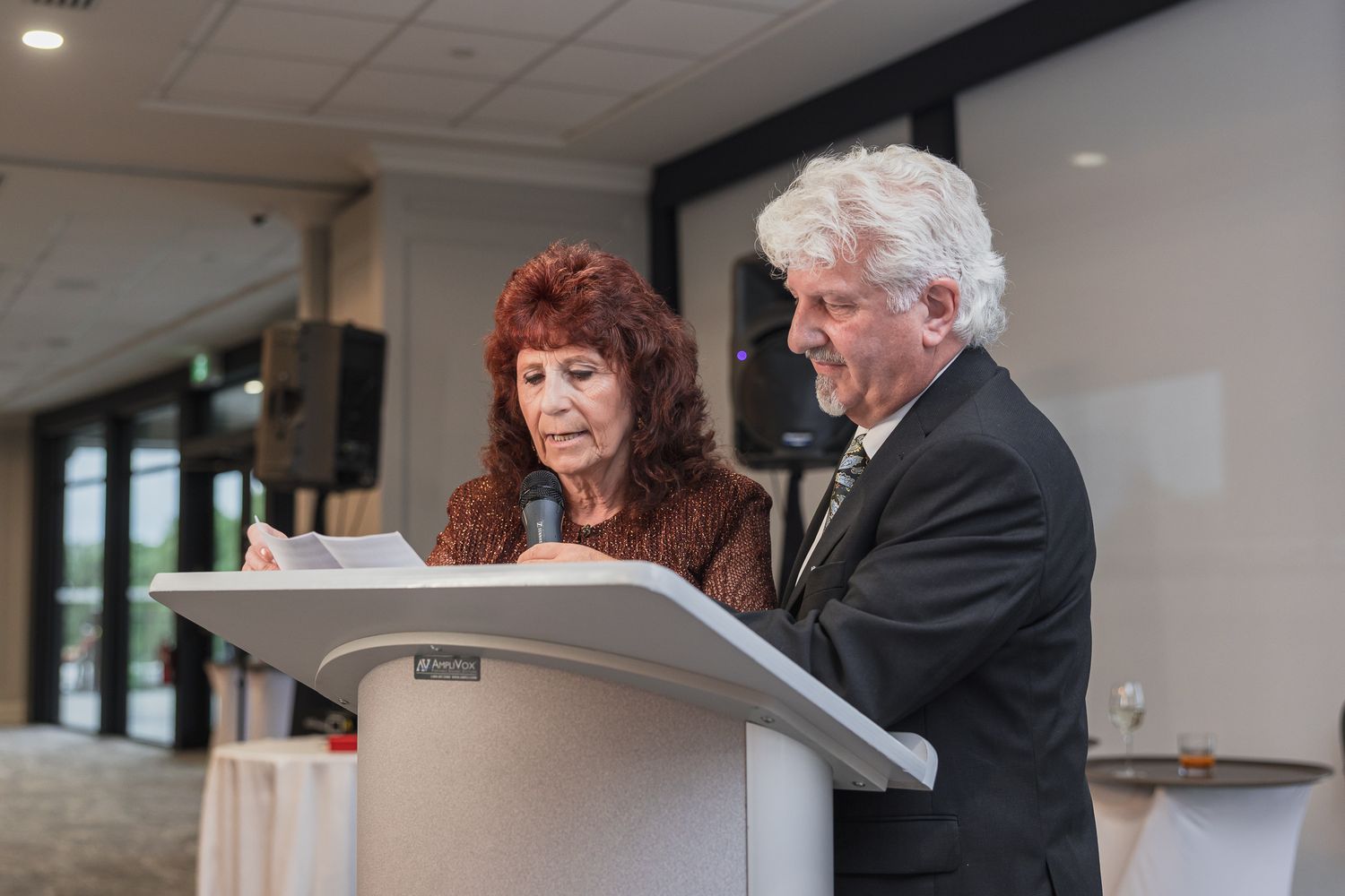 Older couple reads wedding speech together at podium during elegant indoor wedding reception.
