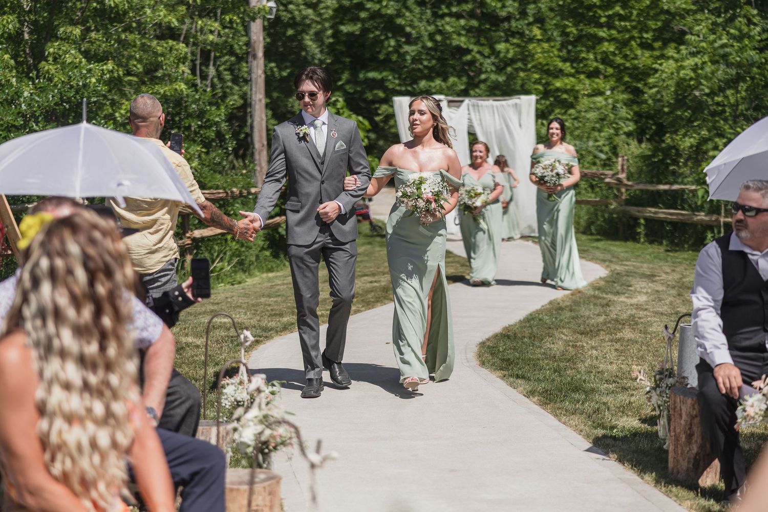 Bride and groom walk down outdoor aisle with bridesmaids in sage green dresses and guests holding umbrellas.
