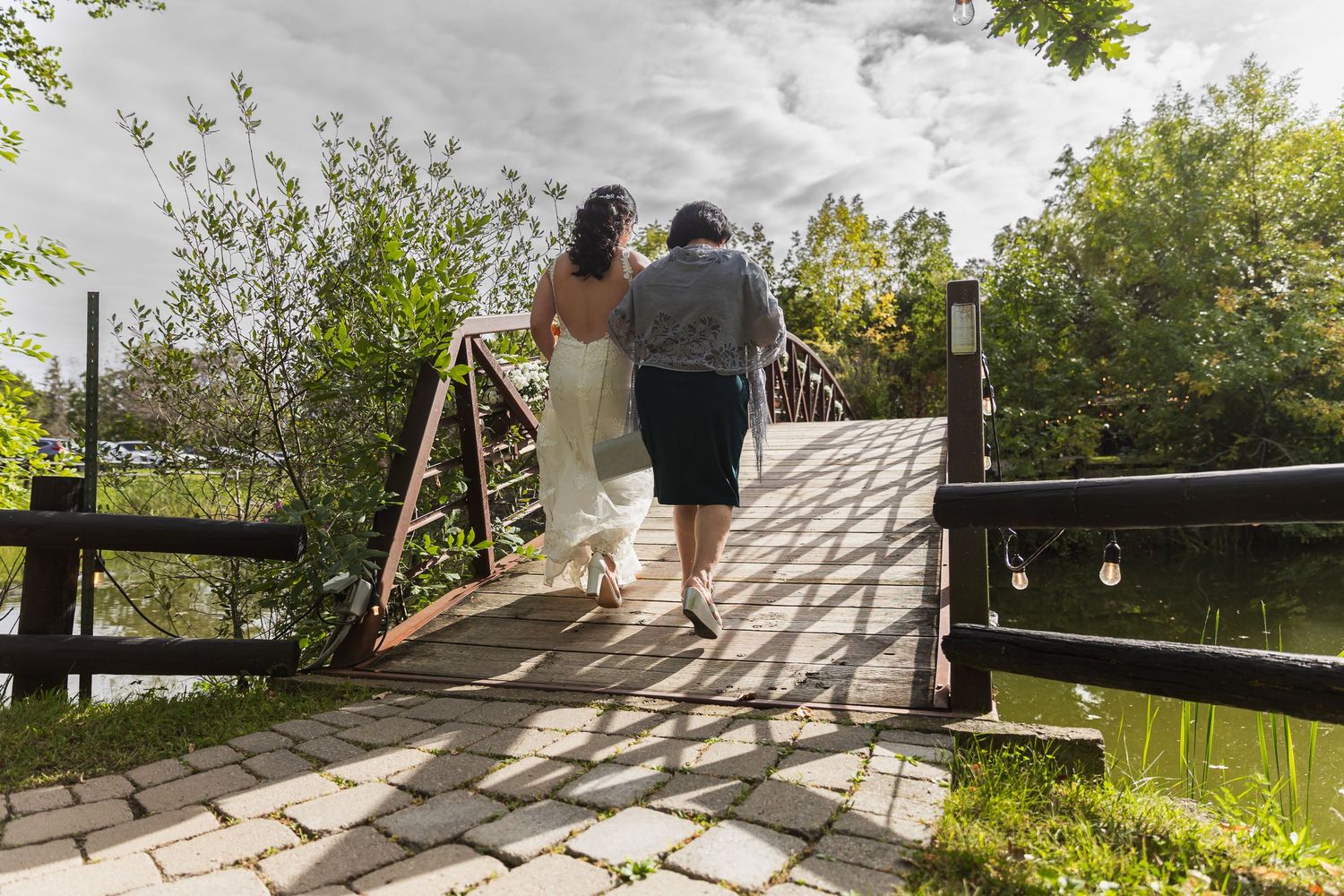 Bride in white gown and companion walk across a wooden bridge in a sunny garden setting.