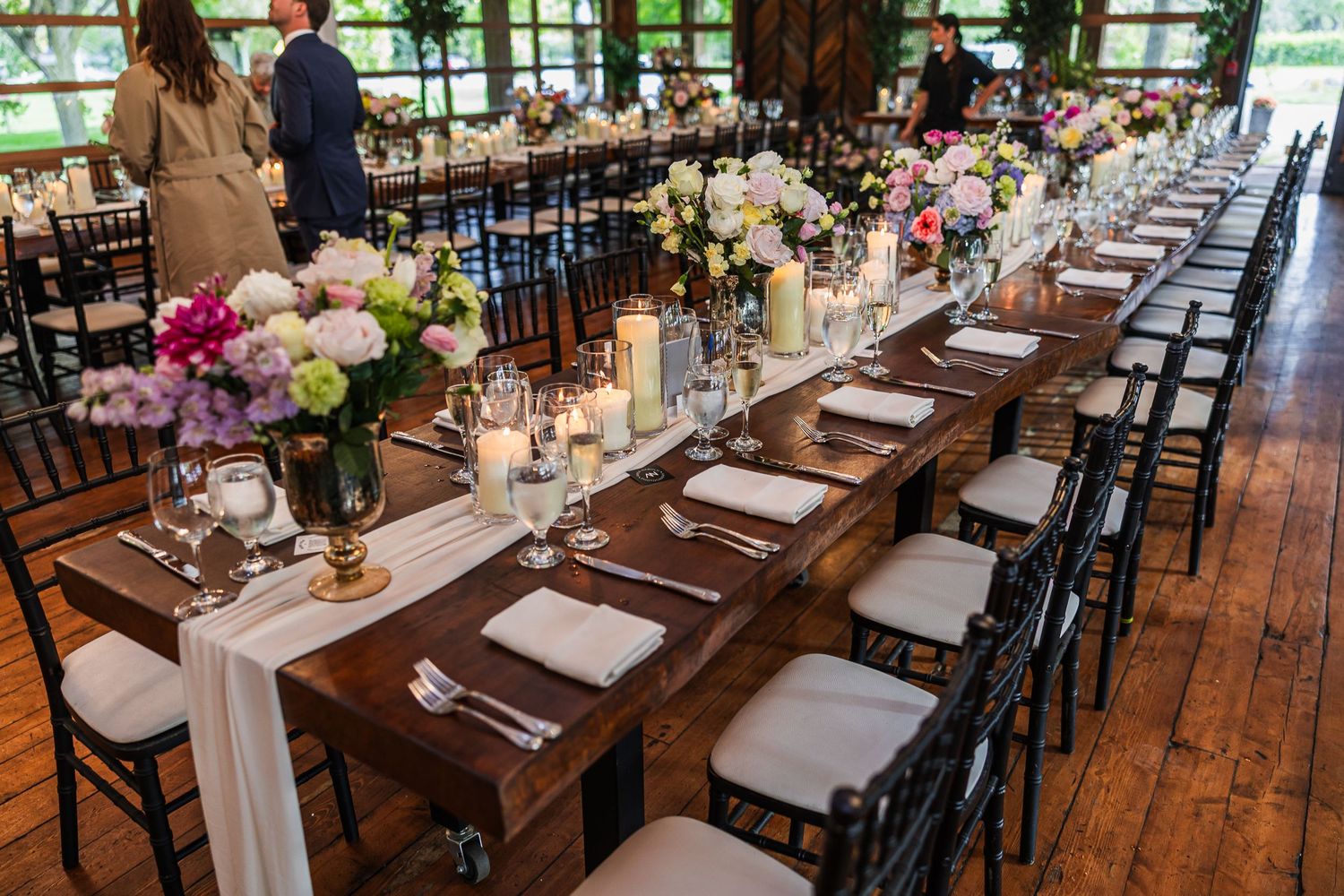 Elegantly set long wooden farm table with floral centerpieces and candles inside a rustic wedding barn.
