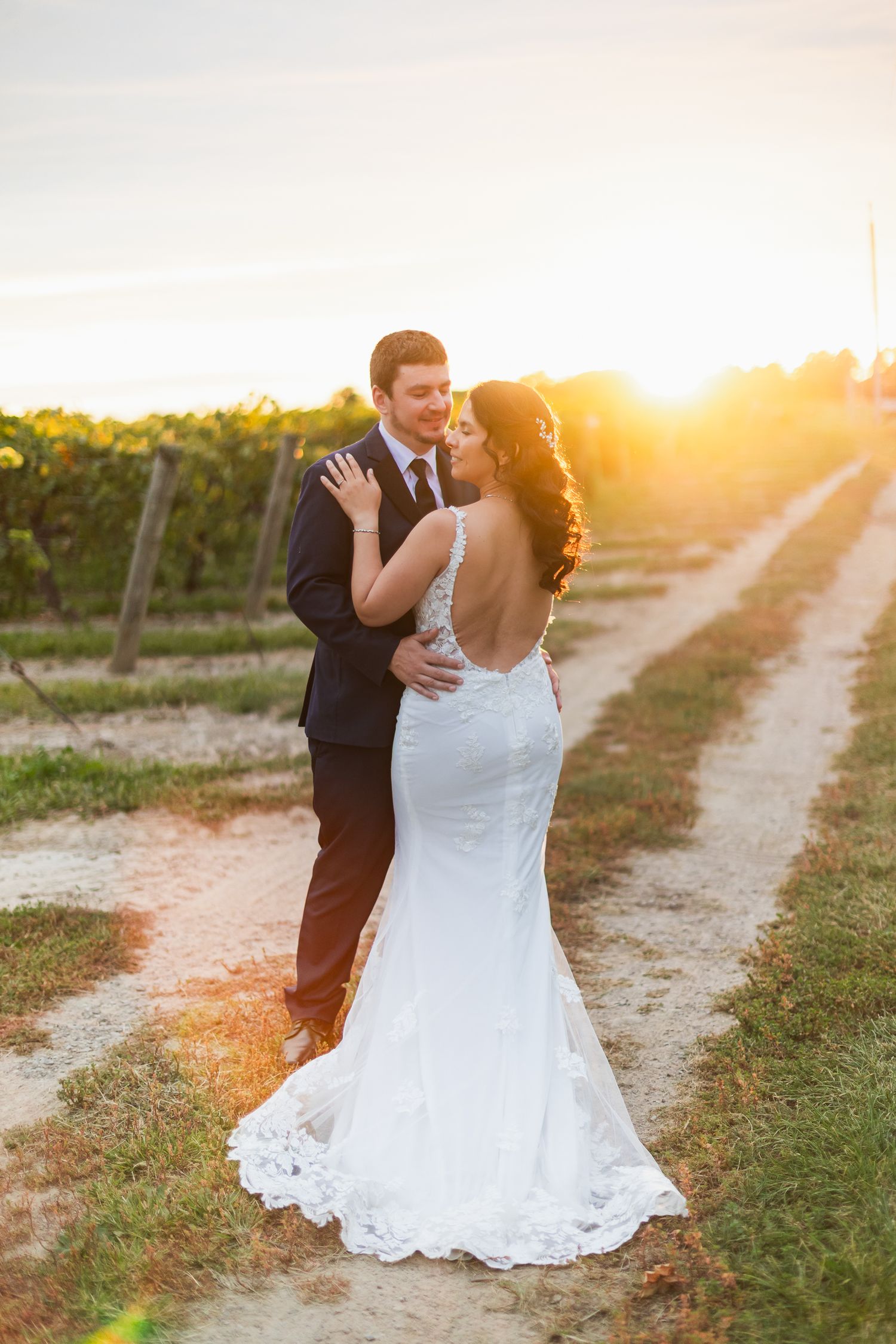 Bride and groom embrace at golden sunset in a vineyard, bride wearing a backless lace mermaid wedding gown.
