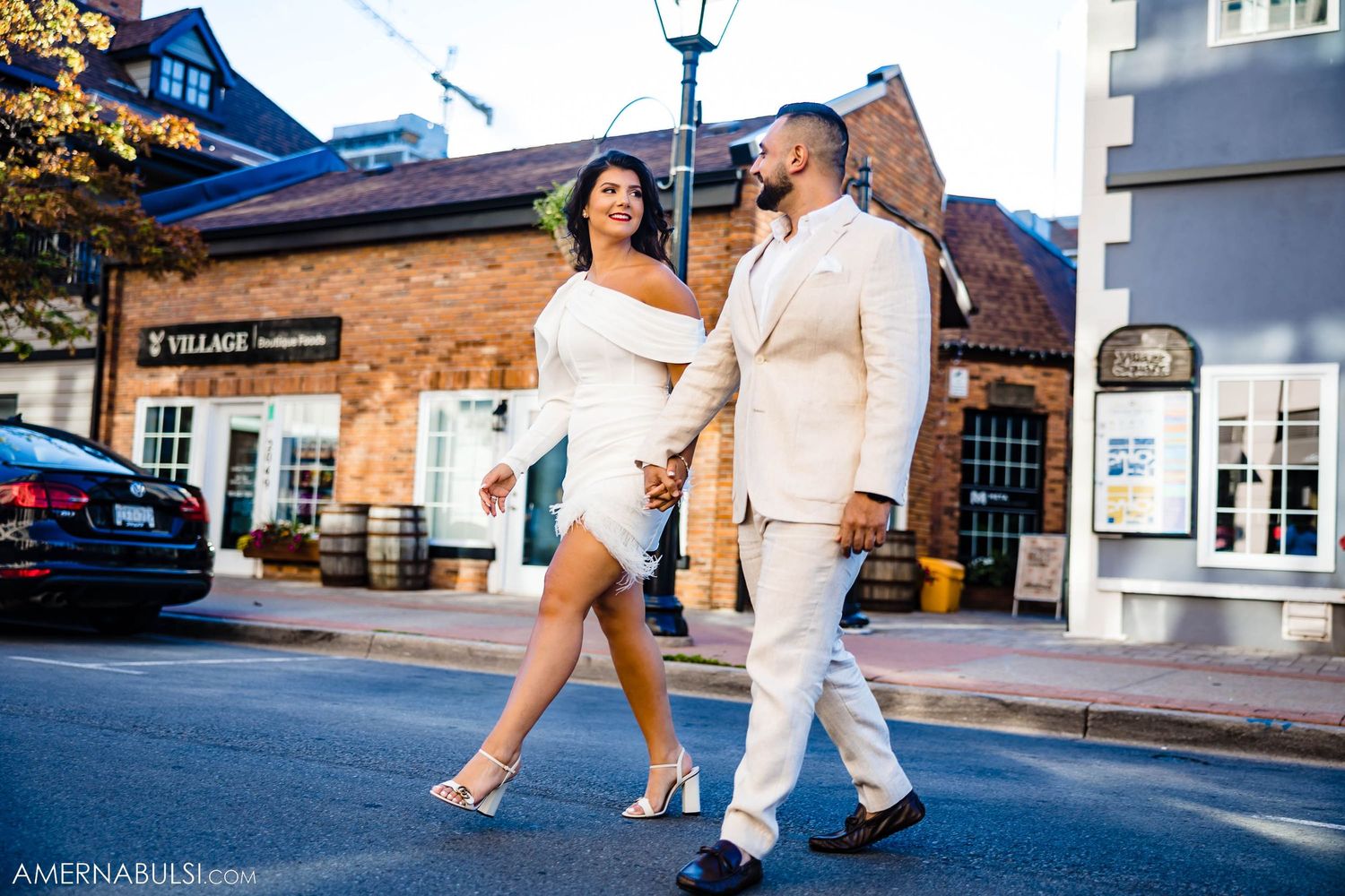 A couple in coordinating white outfits walking together on a city street with brick buildings.