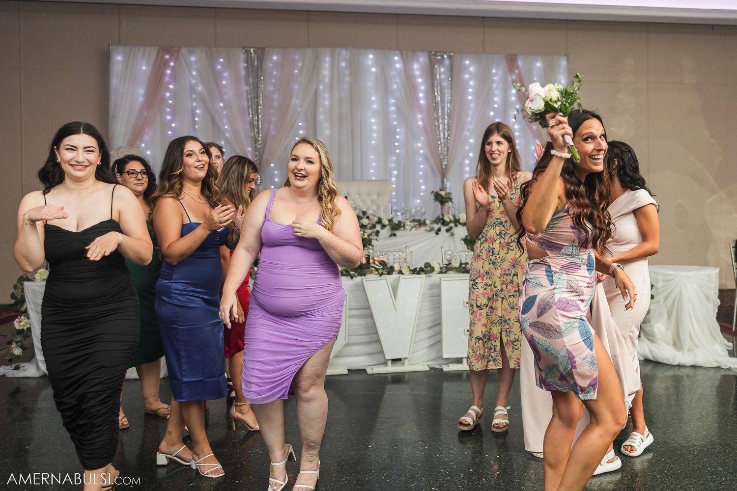 Wedding party members dancing and celebrating at a reception with decorative lights in background.
