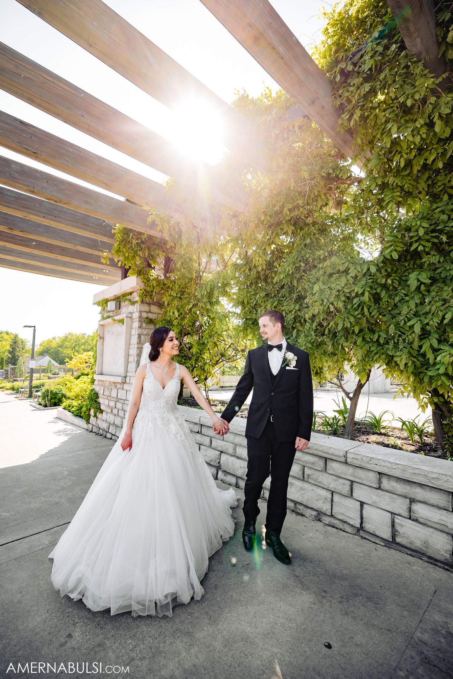 A sunlit wedding portrait under a wooden pergola with climbing vines and stone walls.