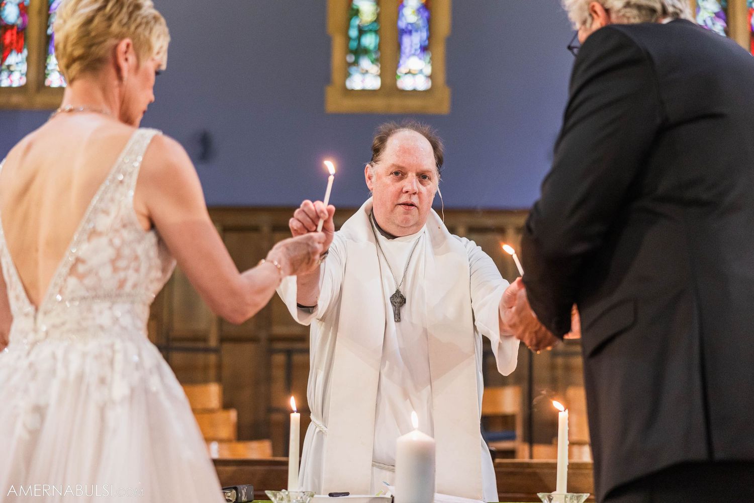 A wedding ceremony takes place in a church as the officiant holds up communion glasses at the altar.
