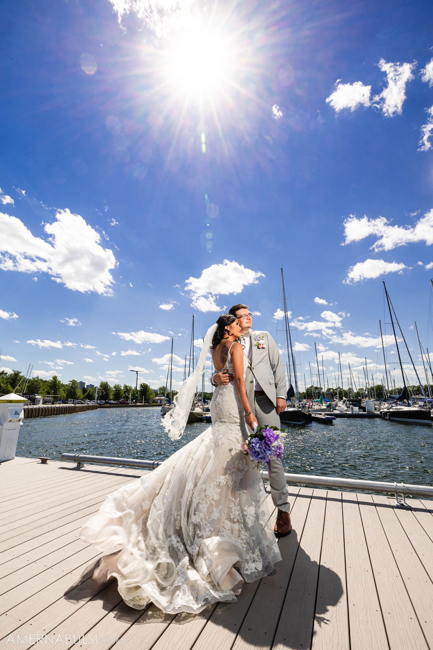 Dramatic sunlit wedding portrait on marina dock with sailboats and bright blue sky.