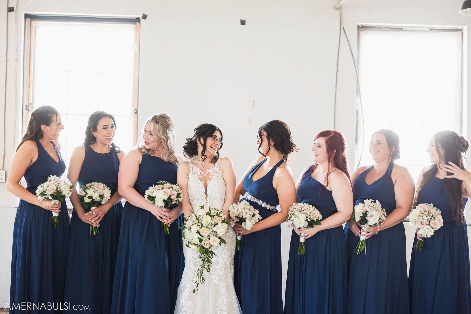Bridesmaids in matching navy blue dresses stand in a row holding white bouquets in a bright indoor venue.
