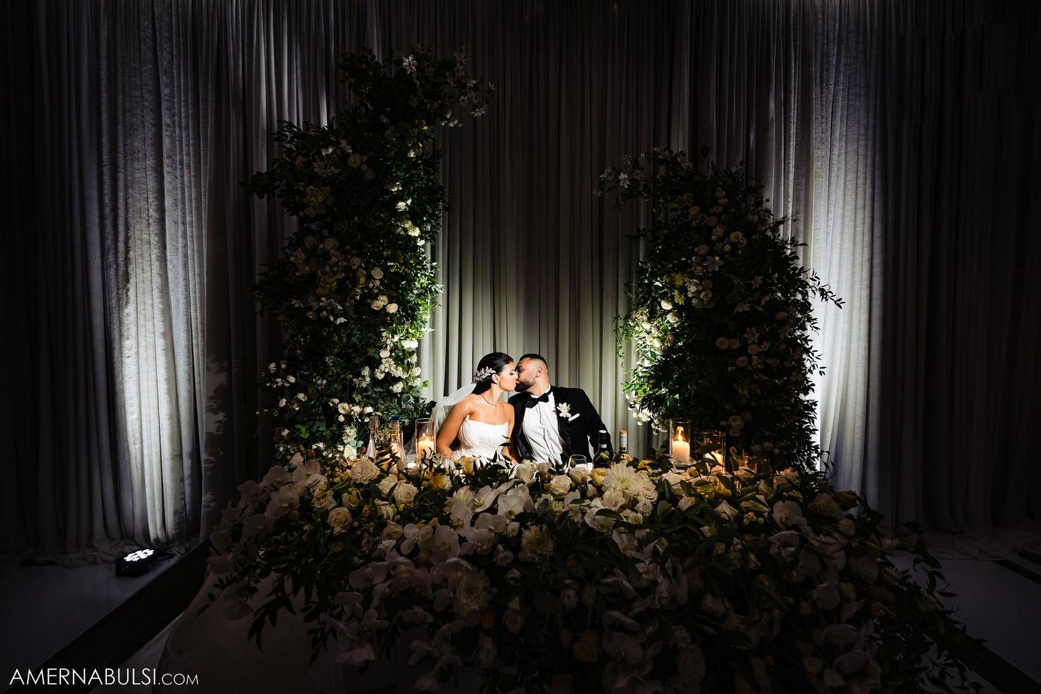 Romantic wedding sweetheart table setup with floral arch and dramatic lighting.