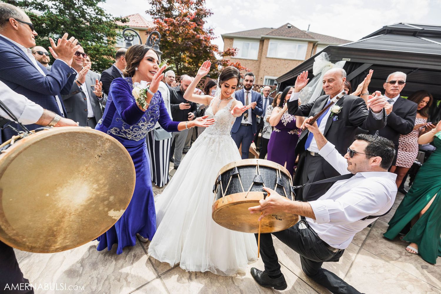 Festive outdoor wedding celebration with musicians playing drums while guests dance and clap.