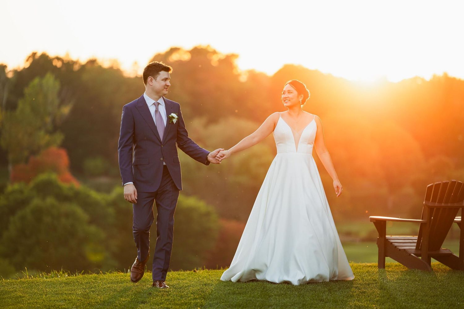 Romantic sunset portrait of newlyweds walking hand in hand on grassy hill.