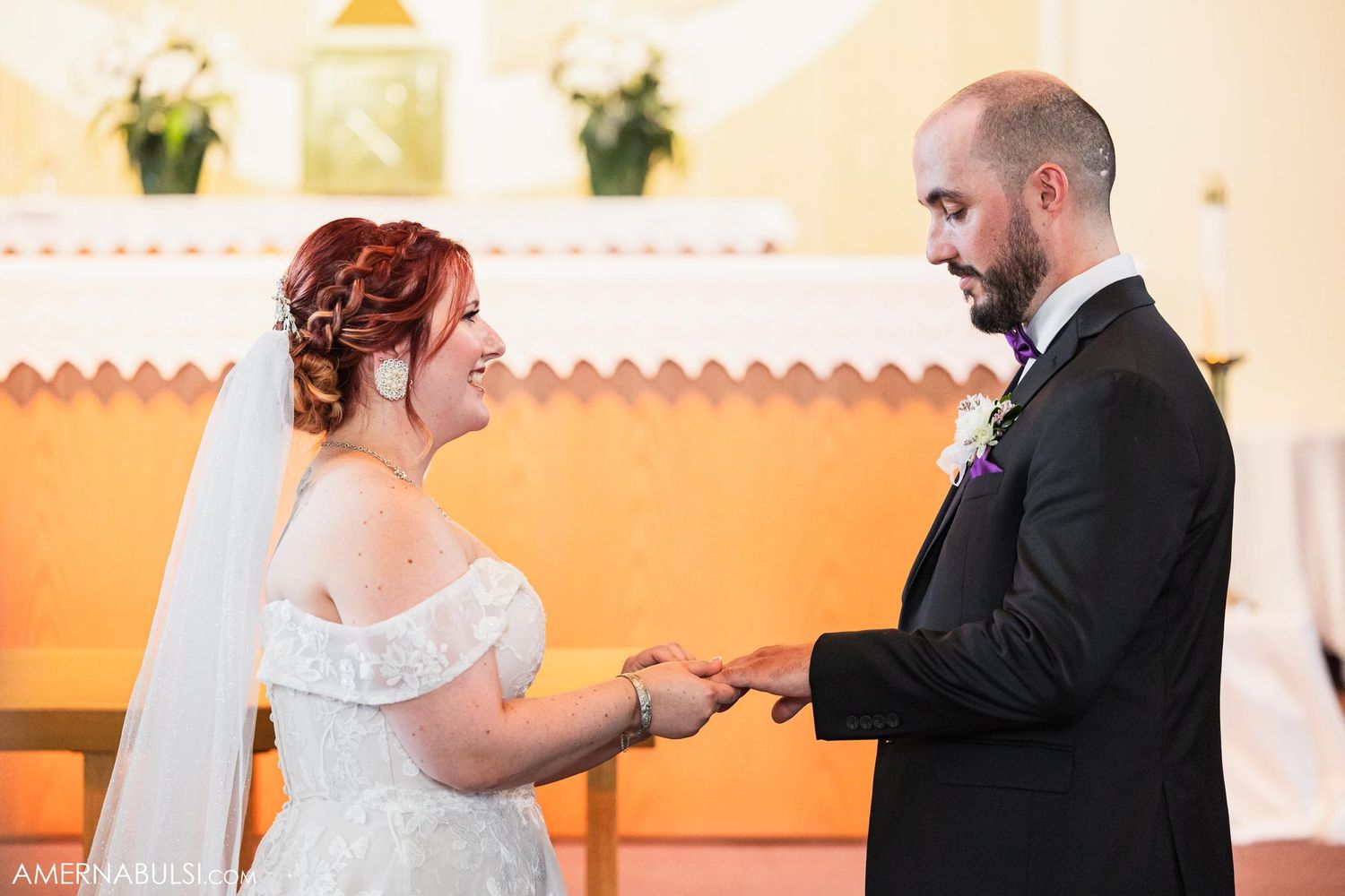 Couple exchanging rings during traditional church wedding ceremony with orange walls and white altar decorations.