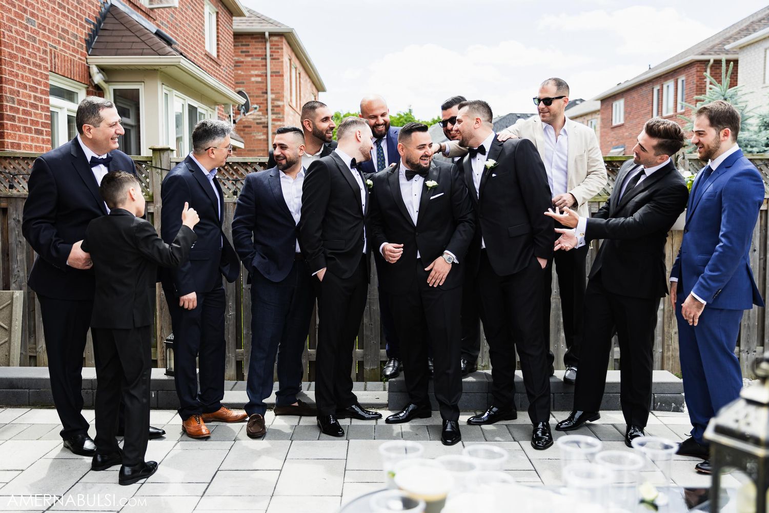 Wedding party in formal attire gathered outdoors for candid group photo against brick building backdrop.