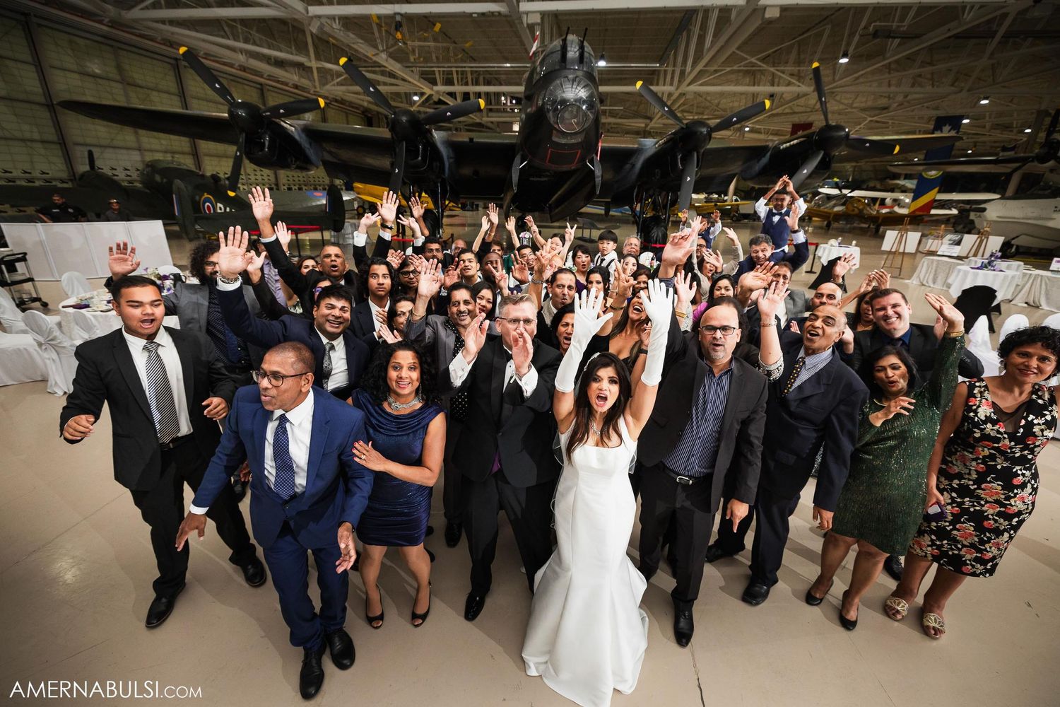 Large group wedding photo with guests and newlyweds cheering in industrial venue setting.