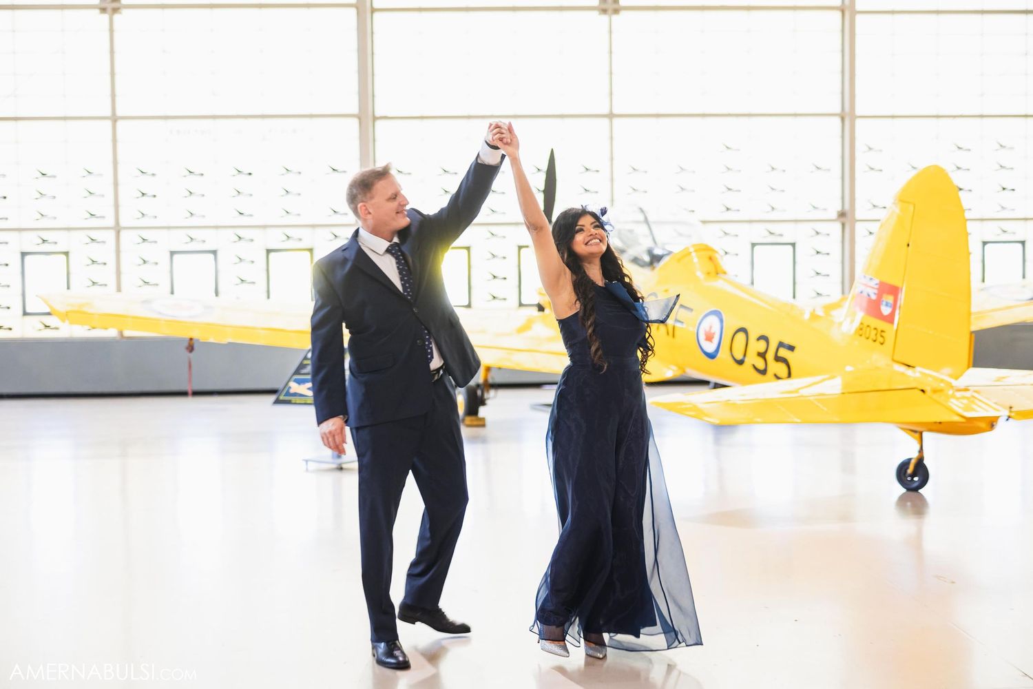 A couple dances joyfully in a hangar with a yellow vintage aircraft behind them.