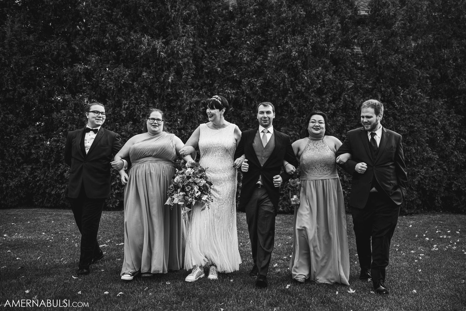 Wedding party in formal attire poses against green hedge backdrop.