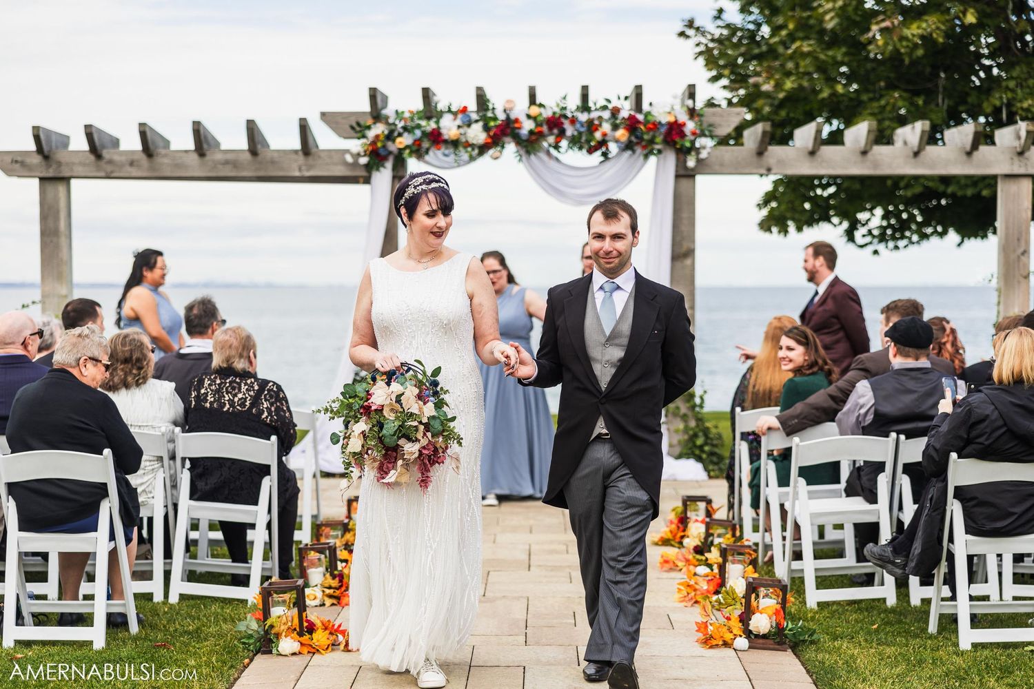 Newlyweds walking down the aisle decorated with autumn pumpkins and flowers at an oceanside venue.