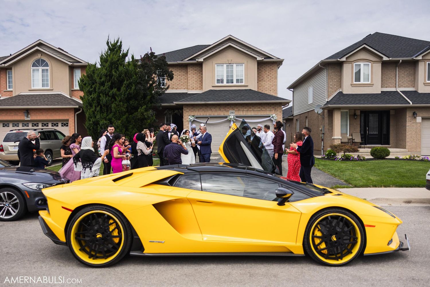 Yellow Lamborghini supercar parked in suburban setting with wedding guests gathering around.