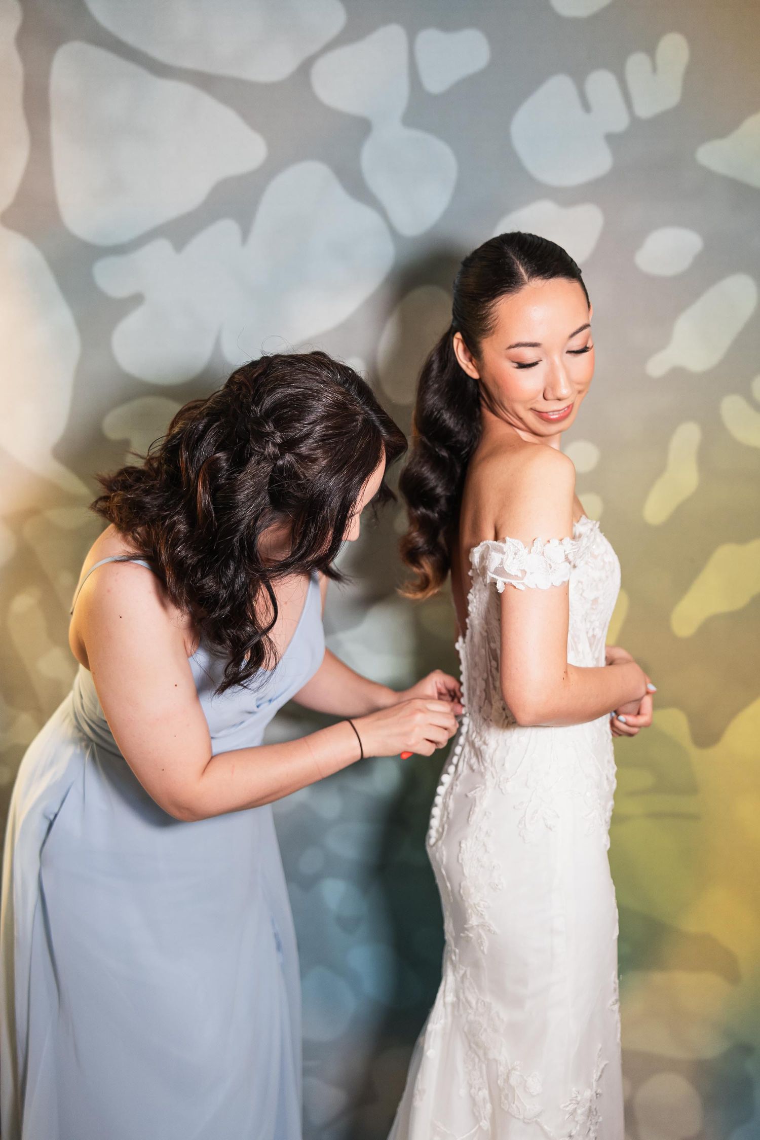 Helper adjusting a bride's fitted lace wedding dress against a decorative wall pattern.