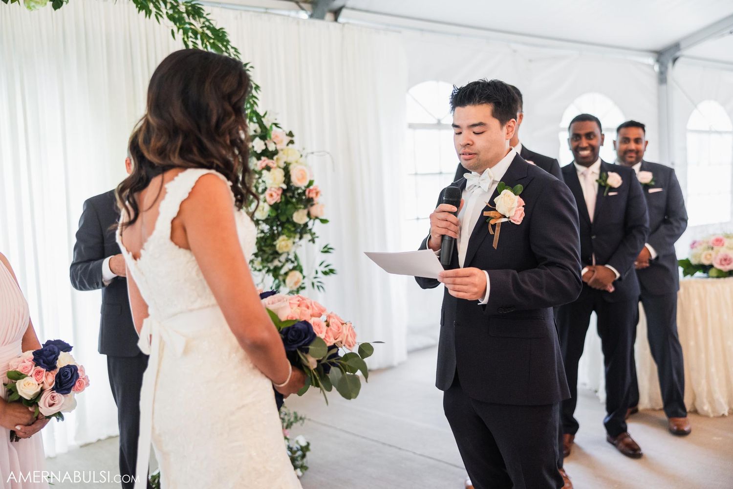 A groom reads his vows during a wedding ceremony in a white tent with floral decorations.