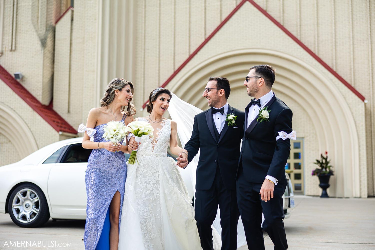 Wedding party laughing together outside church with white car in background during celebration.