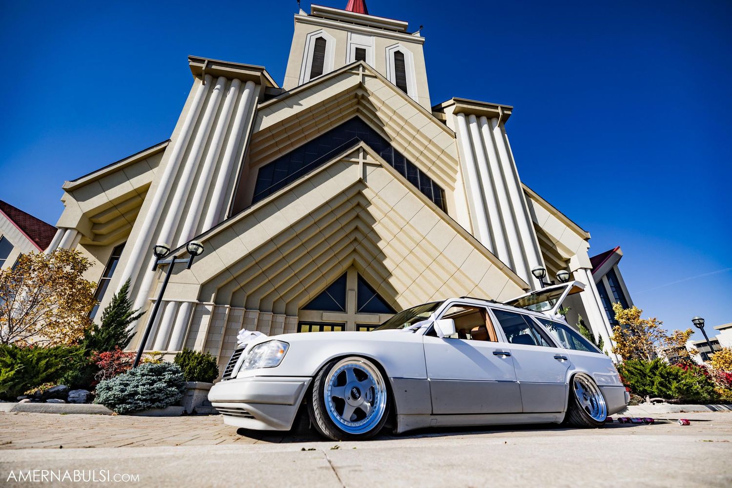 Lowered white custom car parked in front of modern church building with tall spires.