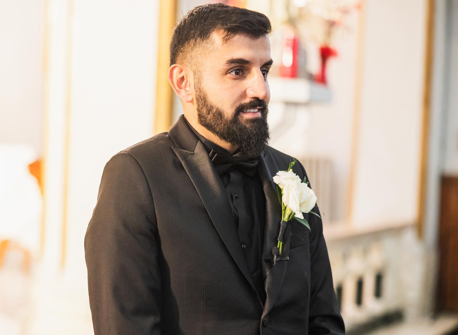 A groom in a black suit with white boutonniere smiles during his wedding ceremony.