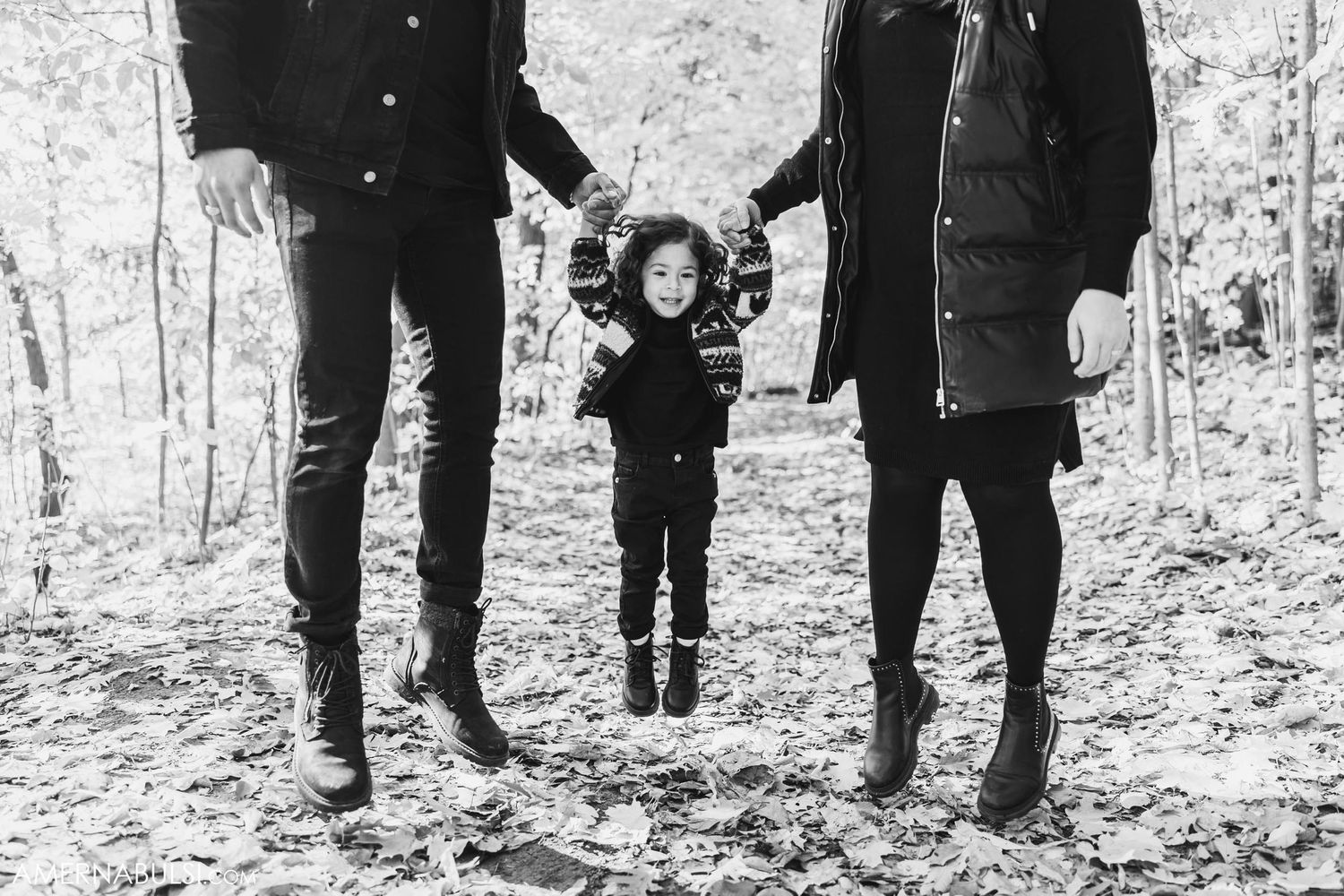 Black and white photo of a jumping child held by adults' hands in a forest setting.