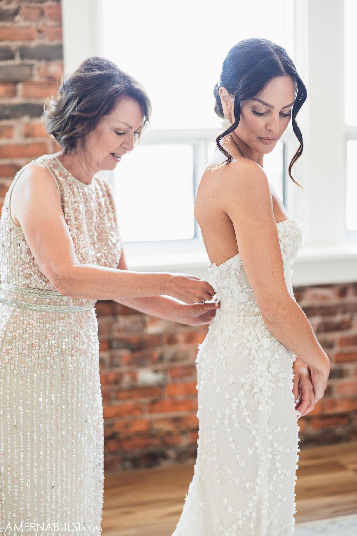 Two women in elegant sequined dresses prepare for a special occasion against a rustic brick wall backdrop.