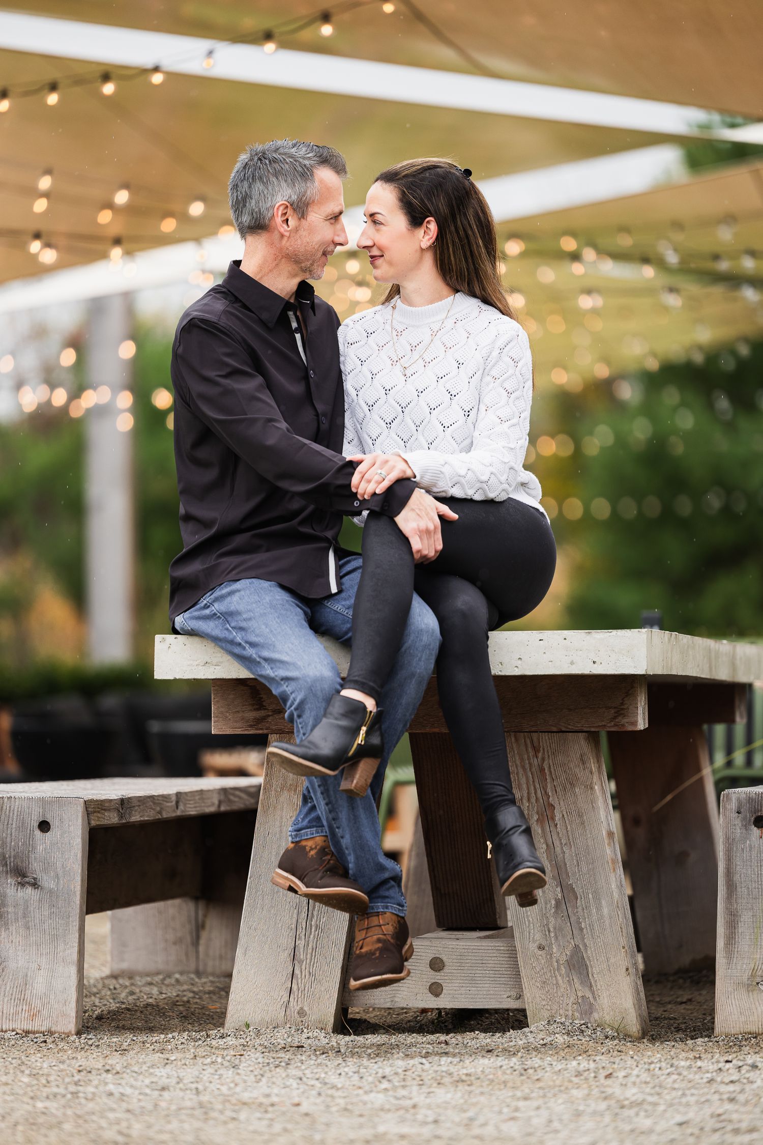 Couple sitting on rustic wooden benches sharing intimate moments under string lights in an outdoor setting.