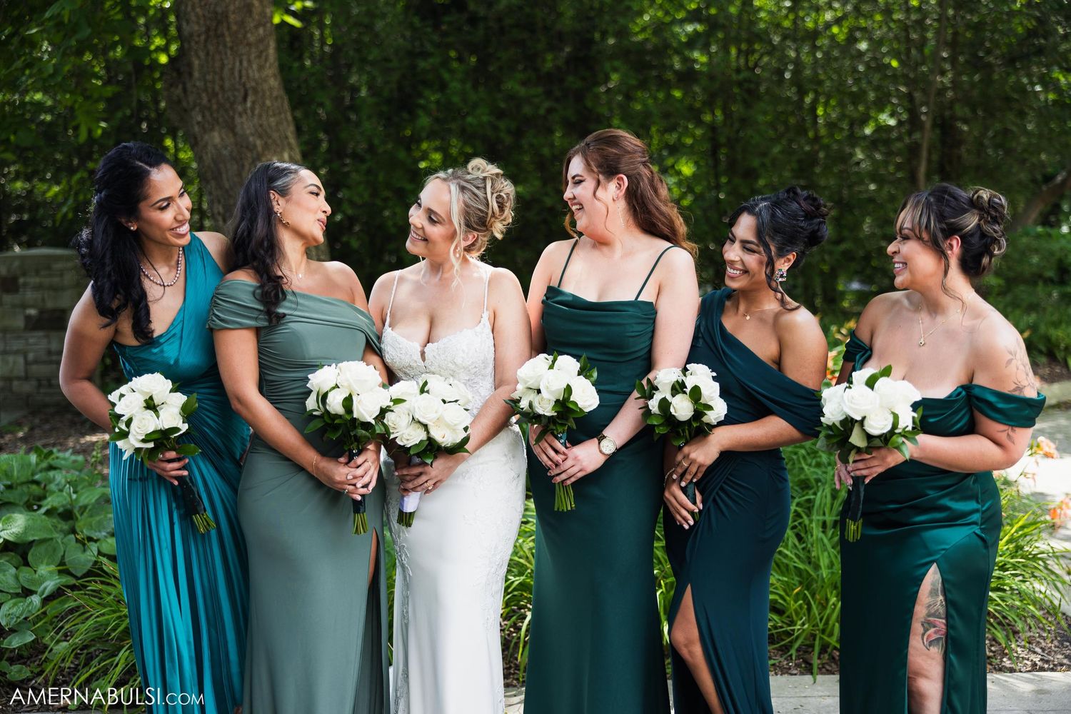 Bridesmaids in varying shades of teal and green dresses laugh together while holding white bouquets outdoors.
