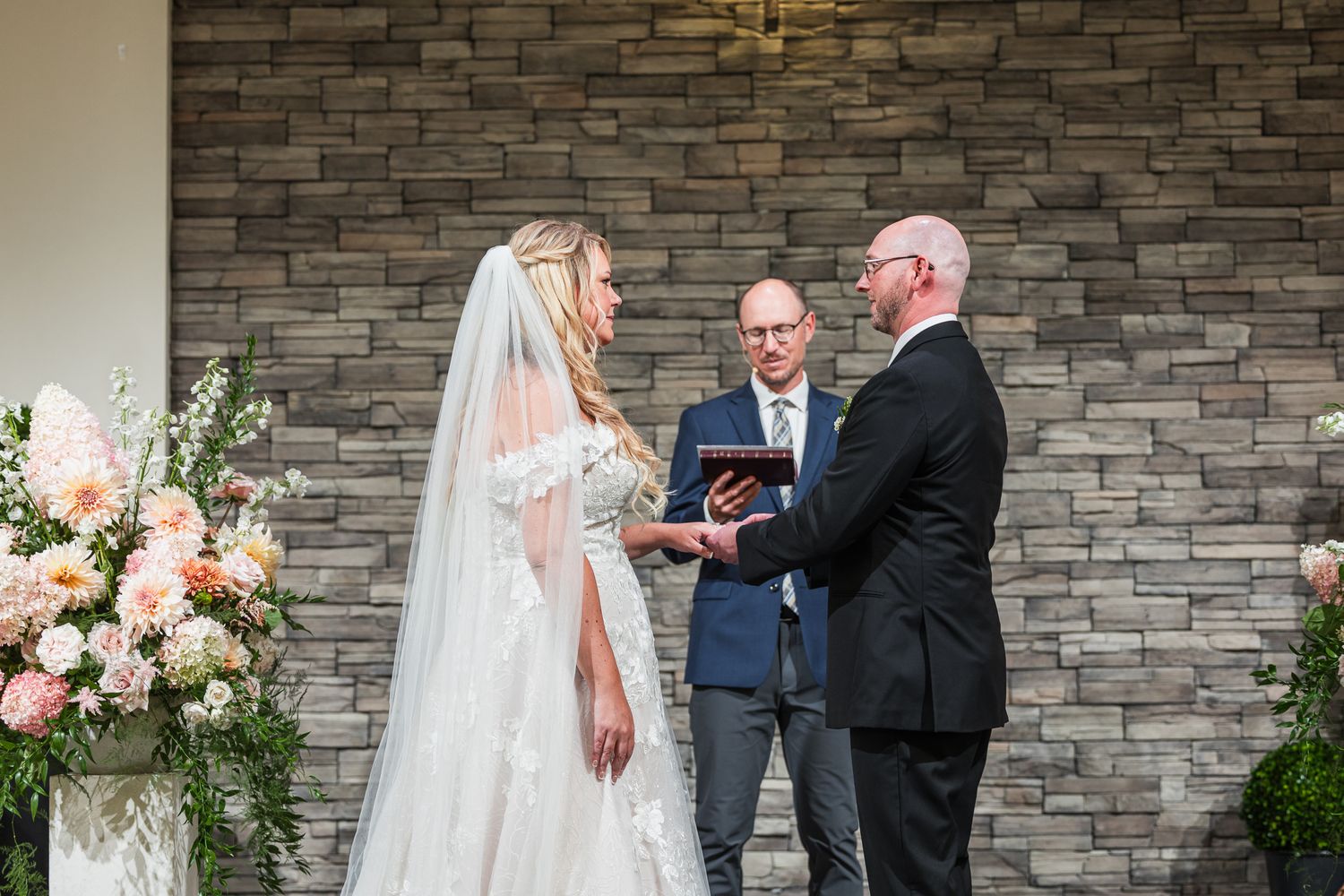 A wedding ceremony takes place outdoors against a stone wall backdrop with pink floral arrangements.