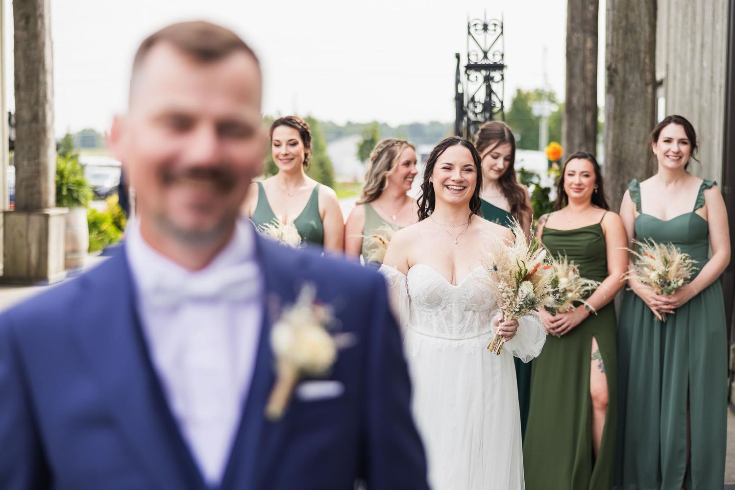 Wedding party photo with focus on groom in blue suit while bridesmaids stand in background.