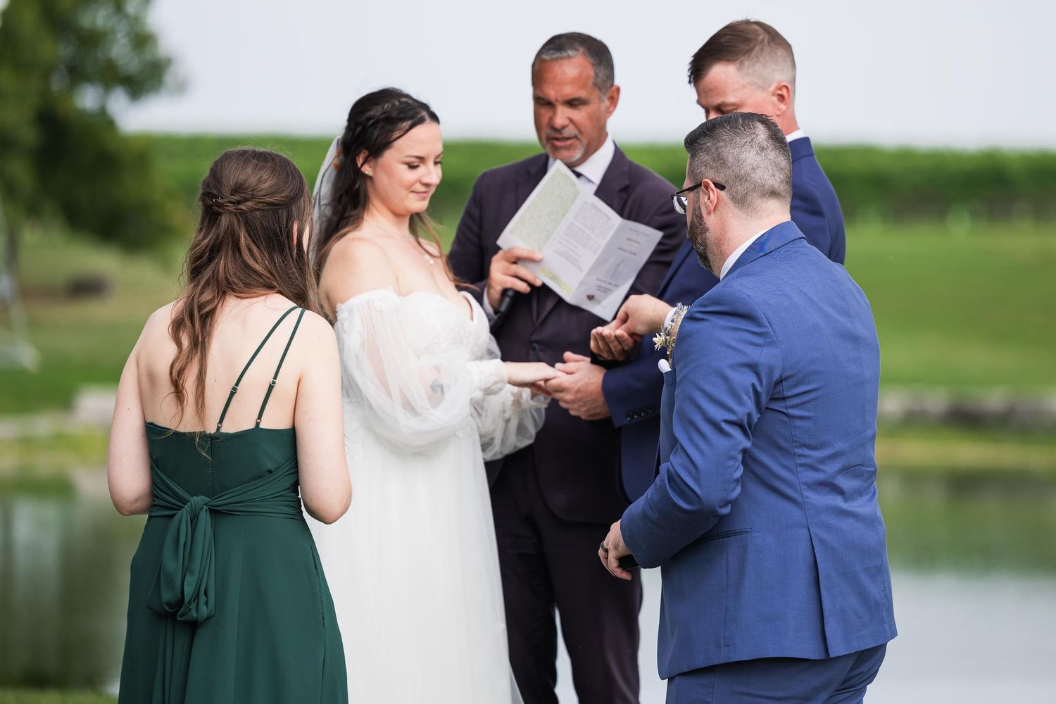 An outdoor wedding ceremony by a lake with attendants in navy and emerald green attire.