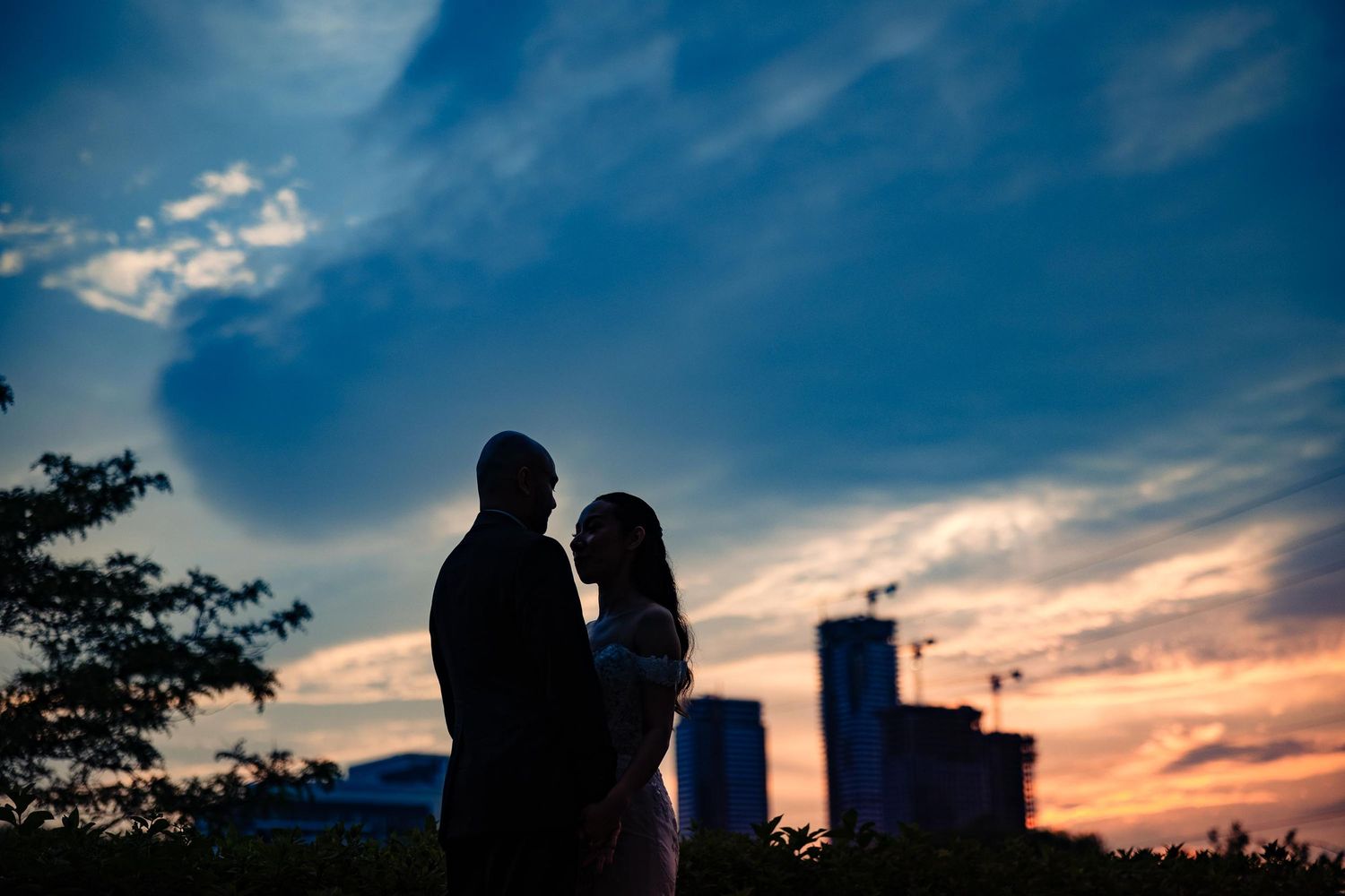 A silhouetted couple stands intimately against a dramatic sunset sky with city buildings in the background.