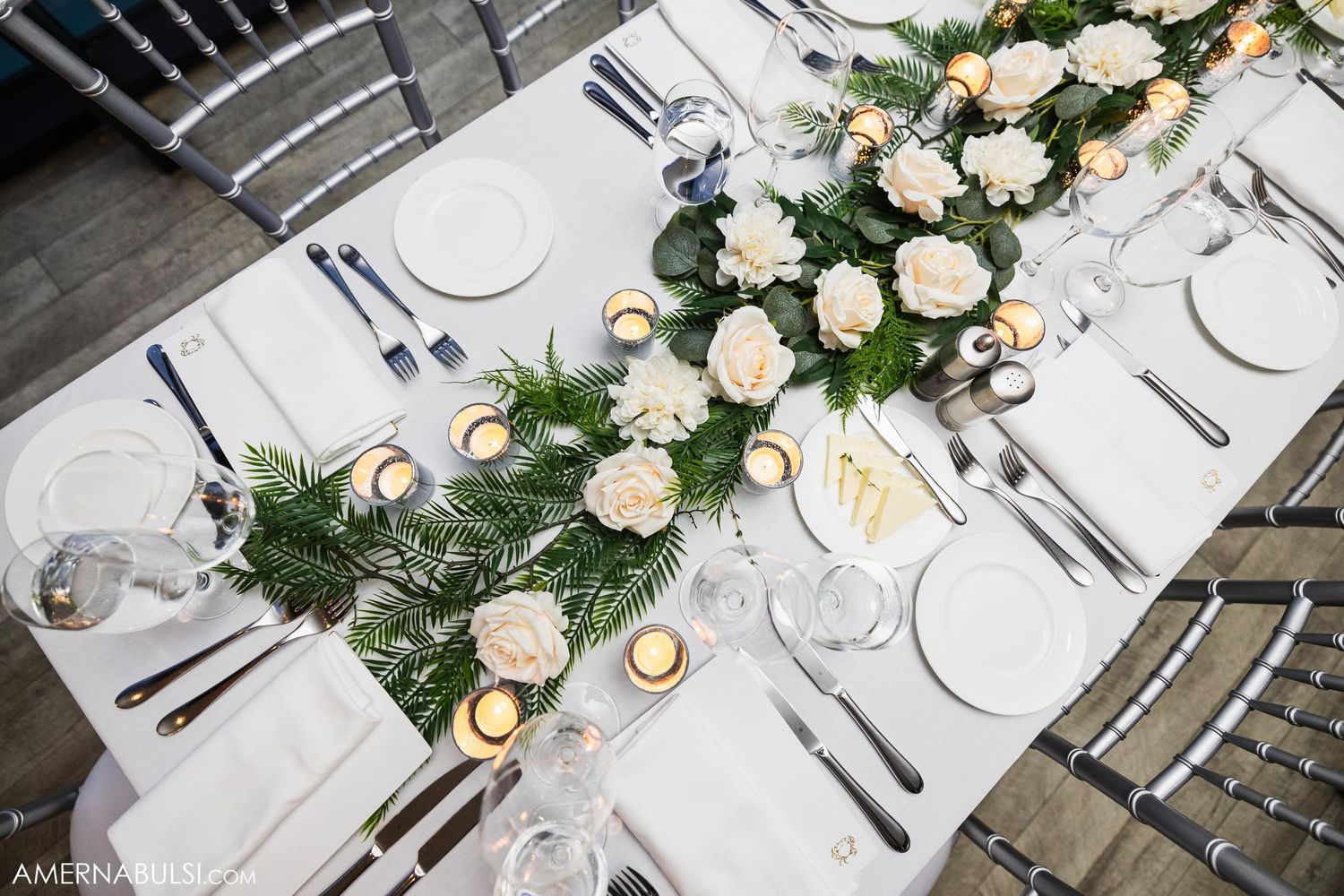 Wedding table decor with white roses and greenery runner on an elegant white tablecloth.