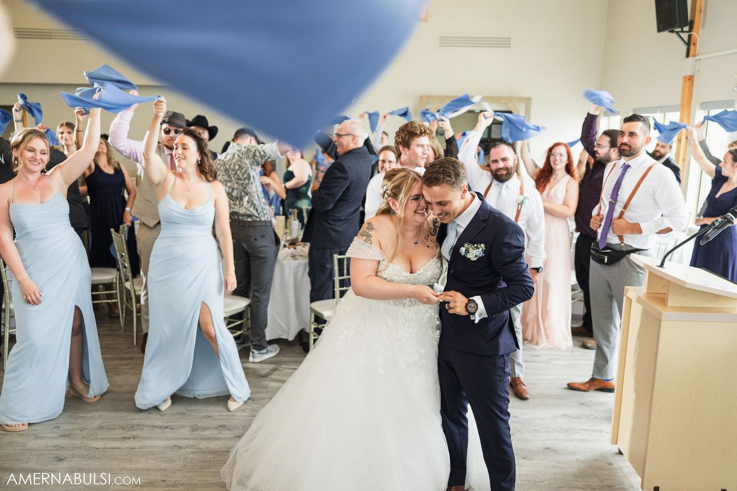 Wedding party guests dance and celebrate in an elegant ballroom with natural light streaming through windows.