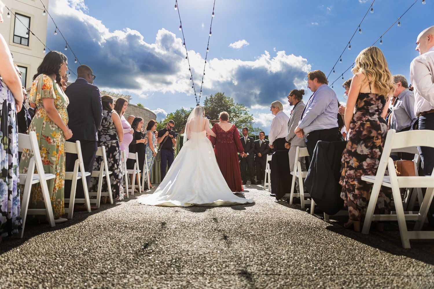 Bride walking down the aisle at an outdoor wedding ceremony with guests seated on white chairs under a blue sky.