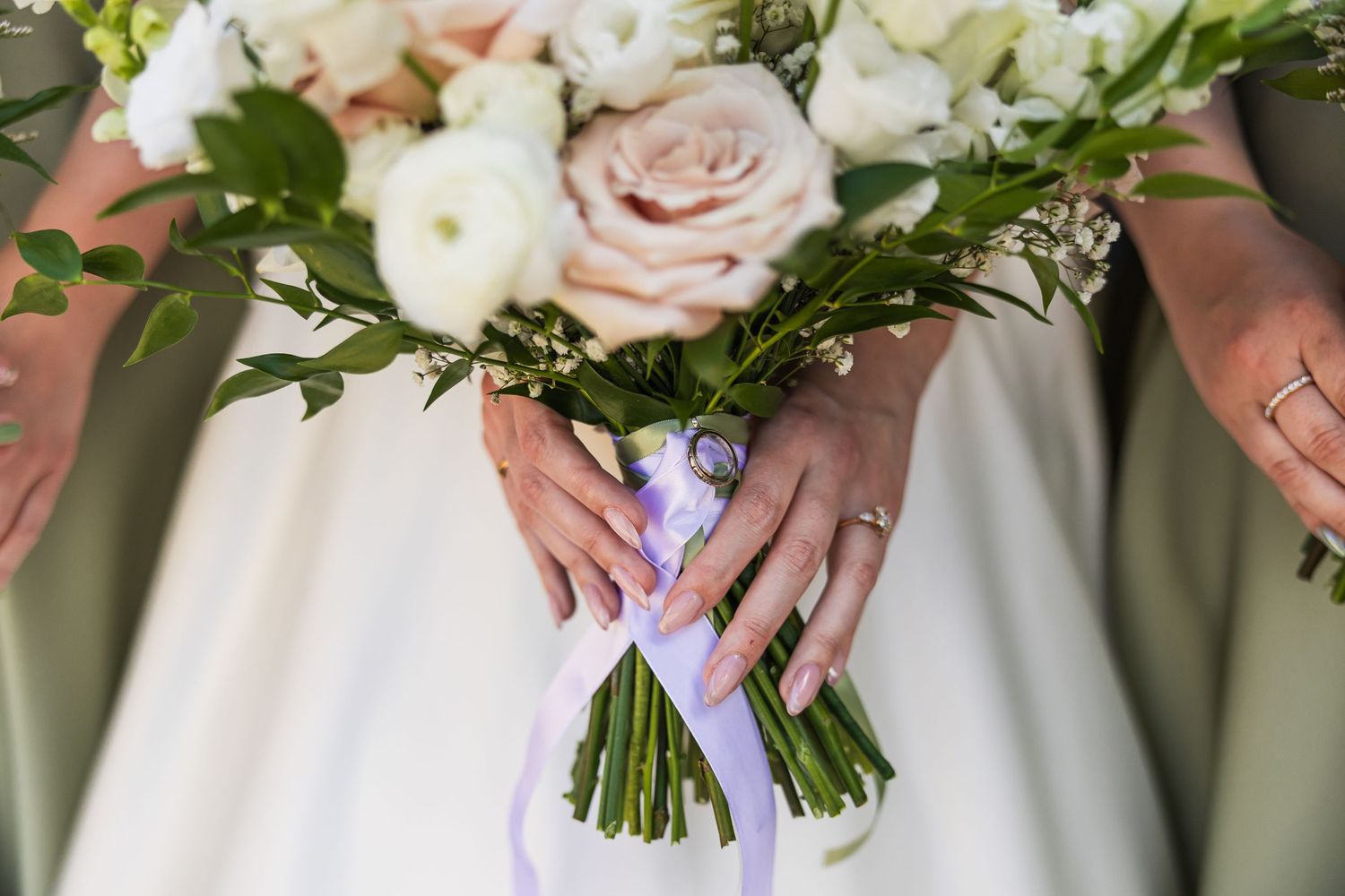 Close-up of bride holding white and blush wedding bouquet tied with lavender ribbon, wedding ring visible.